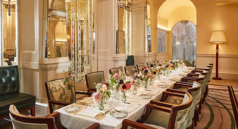 Elegant private dining room at Claridge’s featuring a long table set with white linens, floral centrepieces, and vintage-style chairs beneath mirrored Art Deco arches.