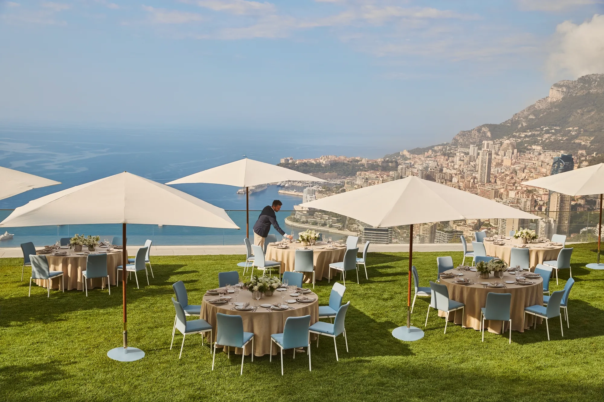 Installation de réception en plein air avec des tables rondes sous de grands parasols blancs sur une pelouse, donnant sur Monaco et la mer, avec un membre du personnel ajustant la mise en place.