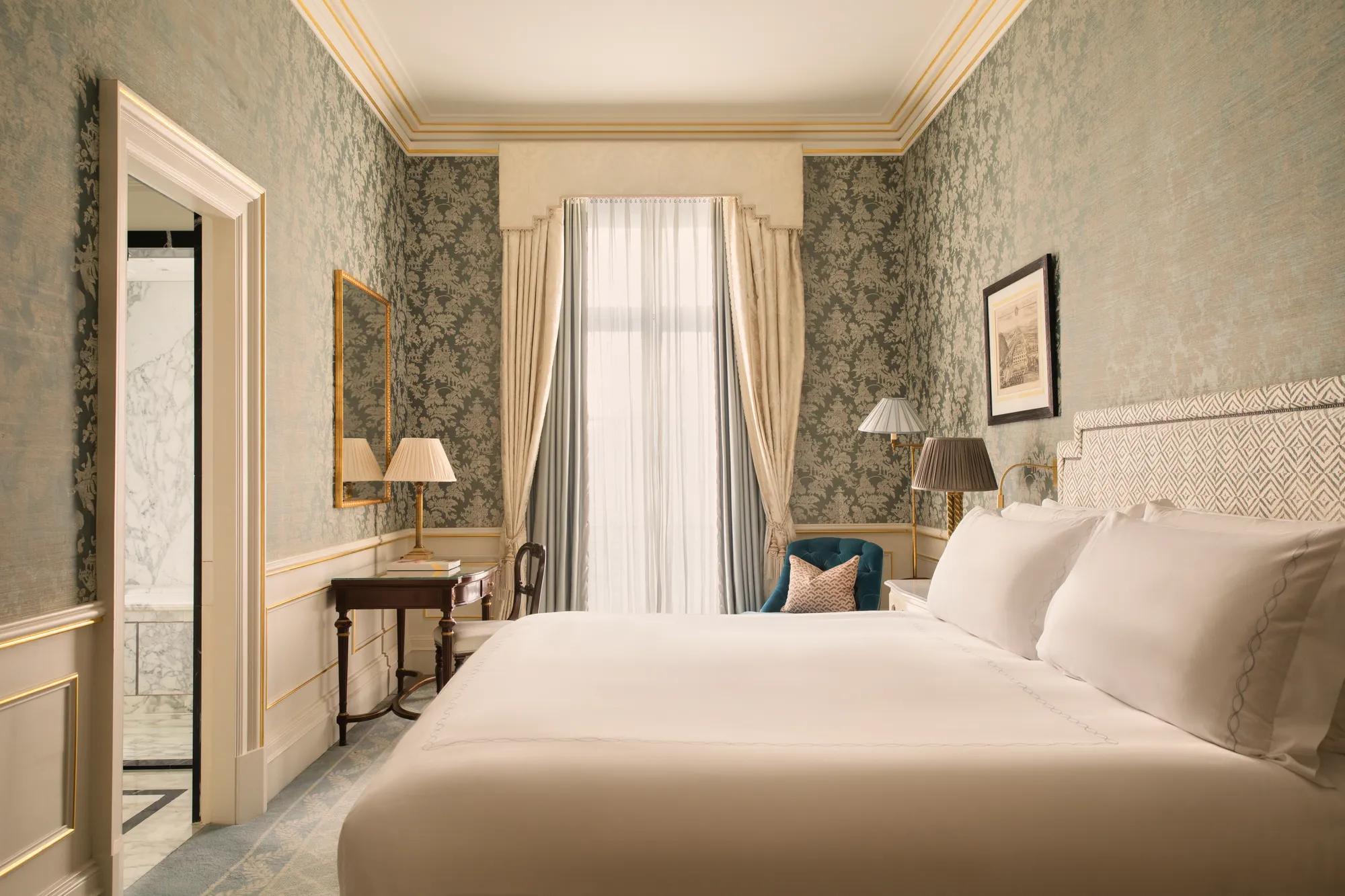 Elegant bedroom with a king bed, blue patterned walls, a writing desk, armchair and tall window with soft daylight.