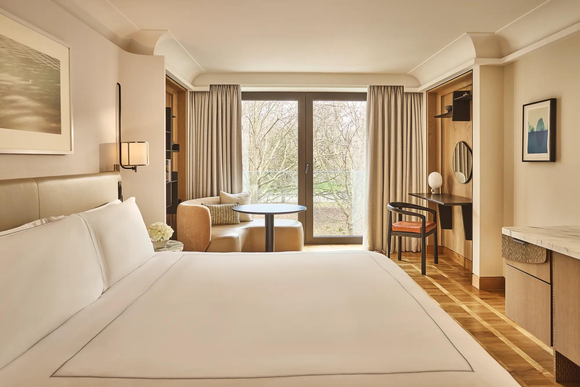 King bed facing balcony doors with park view, armchair and table by window, desk with mirror in warm neutral room.