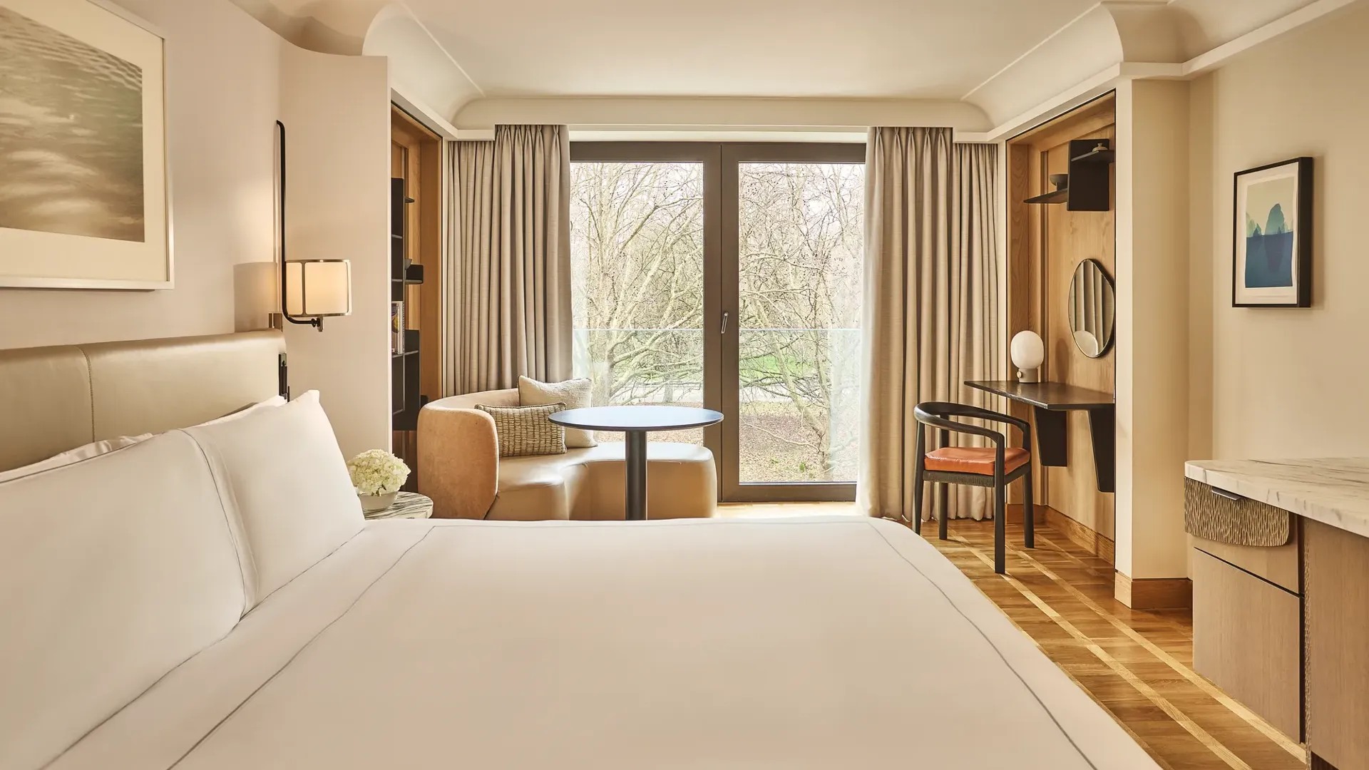 King bed facing balcony doors with park view, armchair and table by window, desk with mirror in warm neutral room.