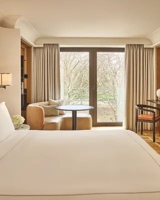 King bed facing balcony doors with park view, armchair and table by window, desk with mirror in warm neutral room.