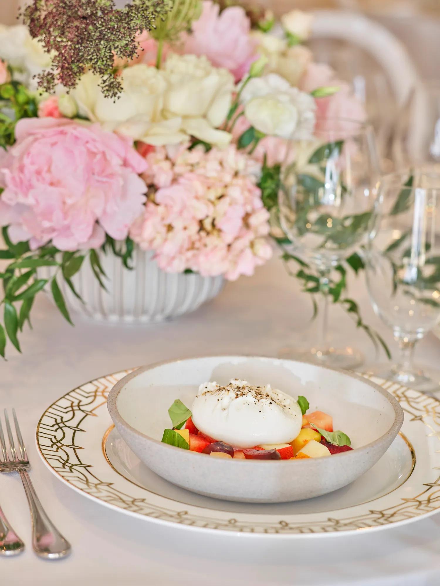 Burrata over summer fruit and basil in a bowl; florals behind.