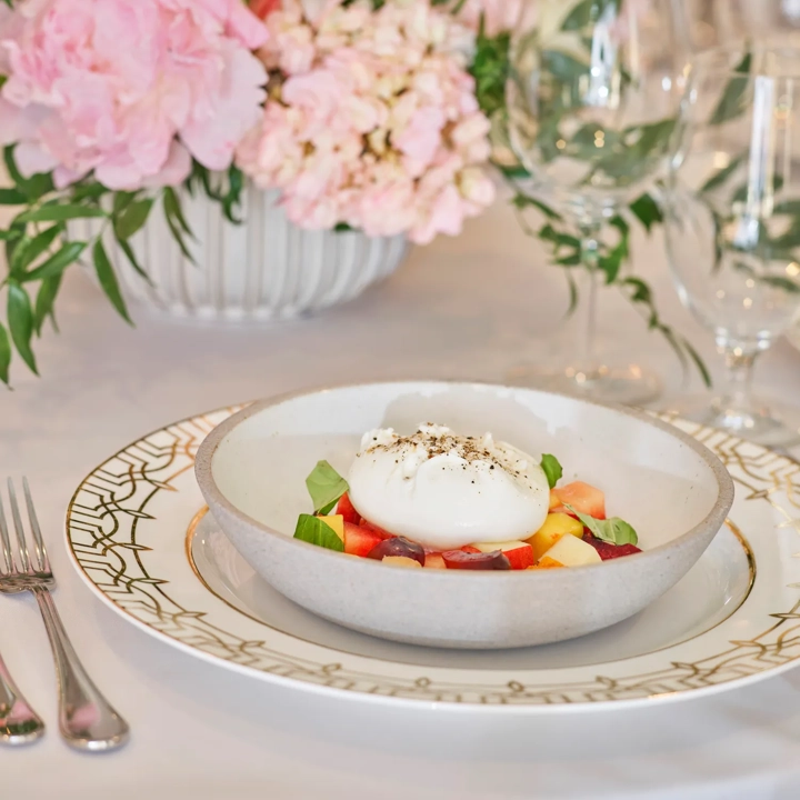 Burrata over summer fruit and basil in a bowl; florals behind.