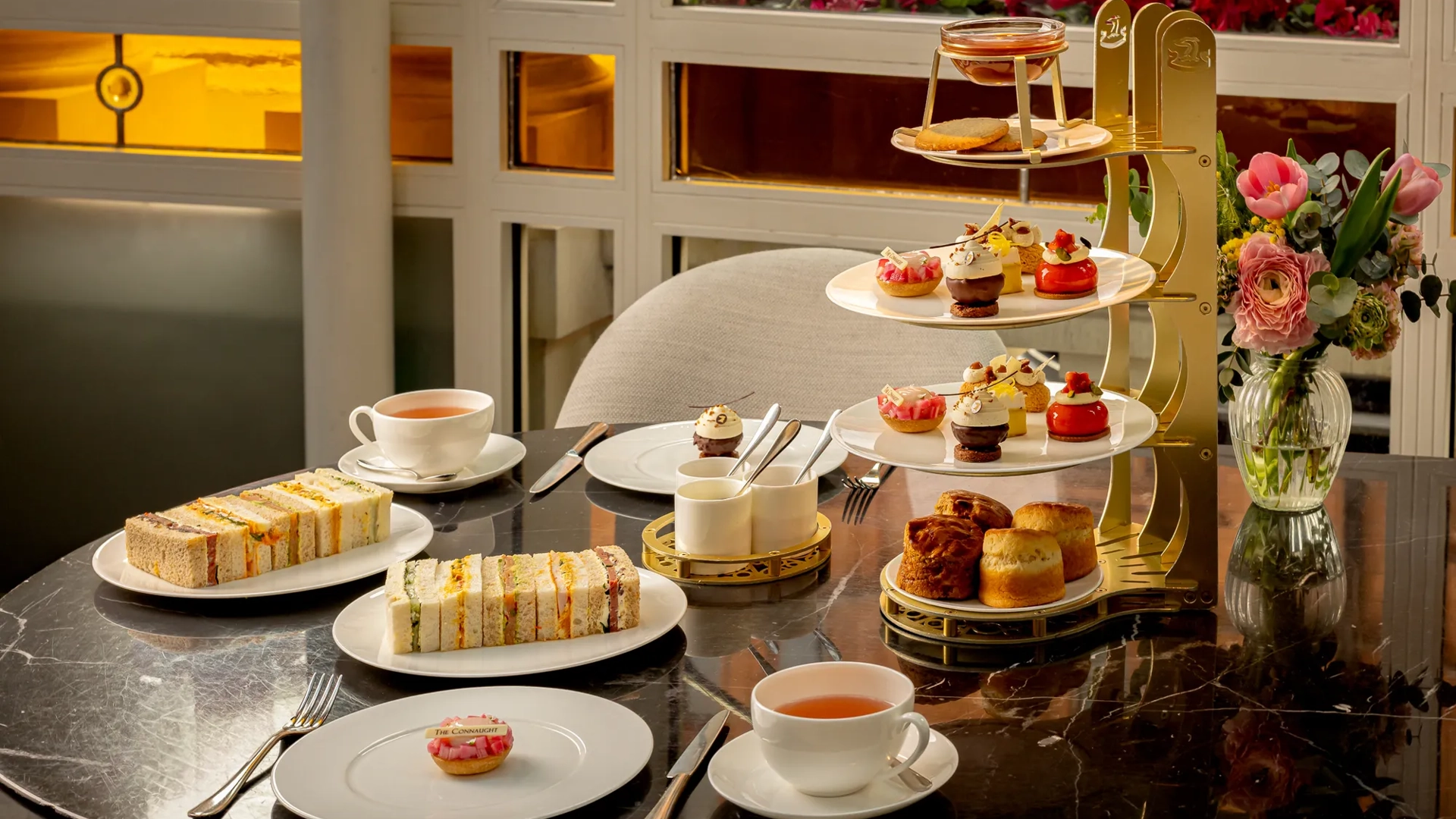 Afternoon tea table with tiered pastries, finger sandwiches, scones, and cups of tea on a marble surface.