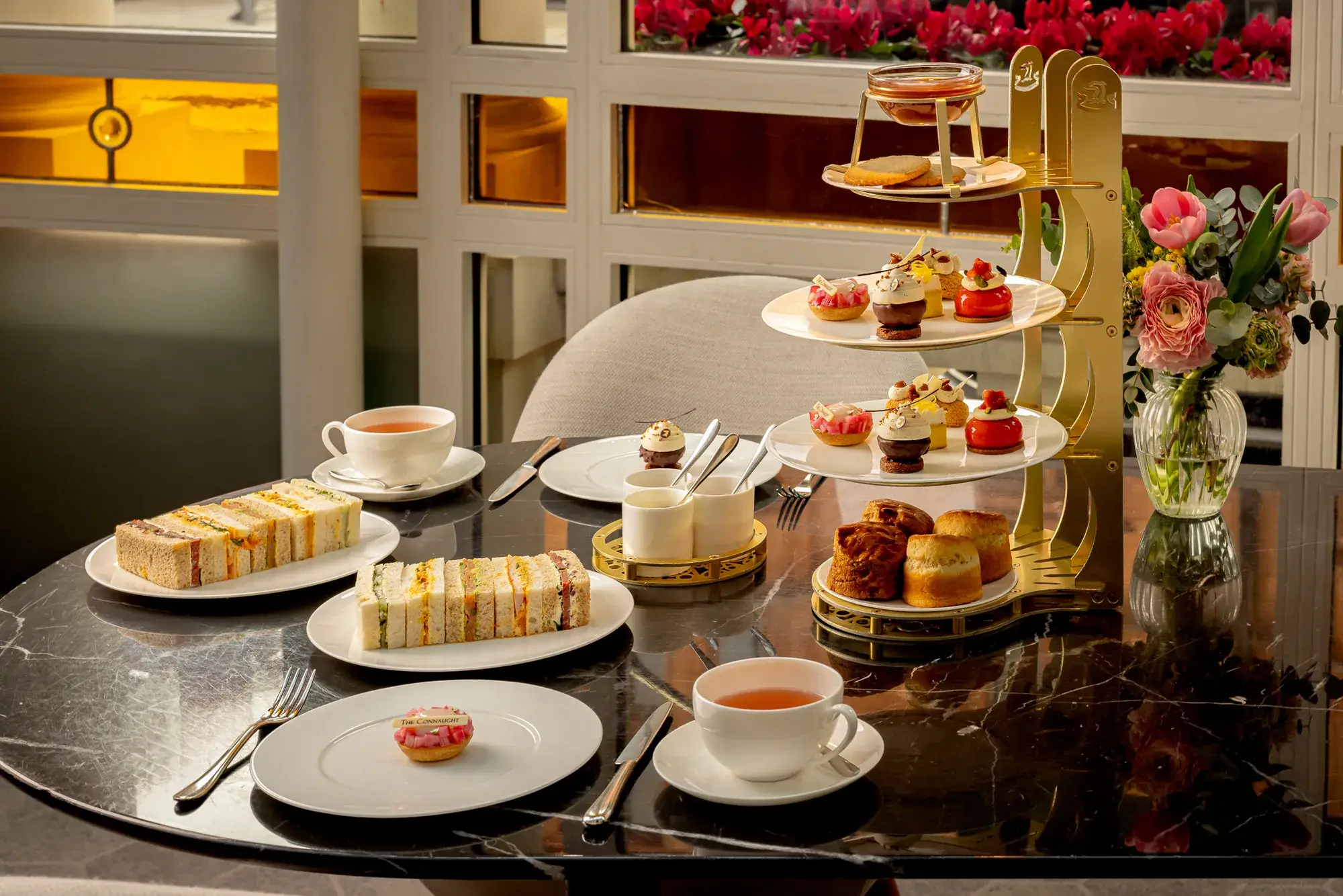 Afternoon tea table with tiered pastries, finger sandwiches, scones, and cups of tea on a marble surface.