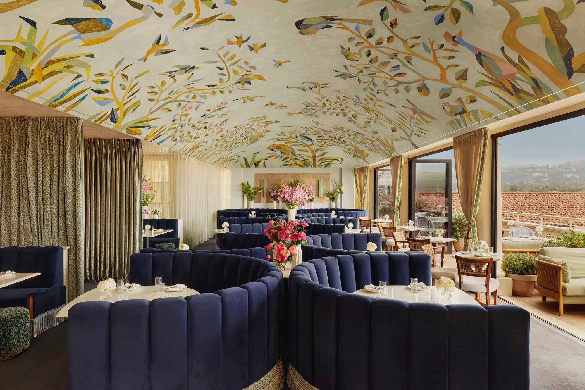 Wide view of Dante Beverly Hills dining room—mural ceiling, curved navy booths, and doors opening to a sunlit terrace.