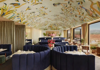 Wide view of Dante Beverly Hills dining room—mural ceiling, curved navy booths, and doors opening to a sunlit terrace.