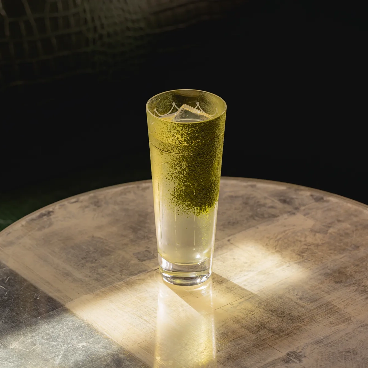 A tall clear cocktail with a green powder rim sits on a sunlit metallic table against a dark upholstered background.