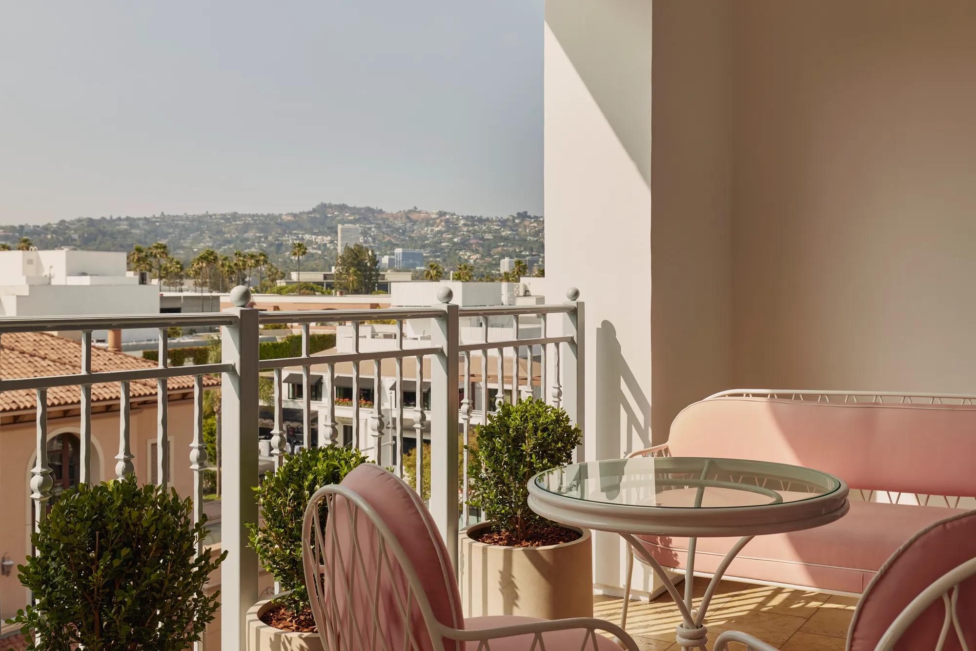 Balcony view with pink cushioned chairs and sofa, glass-topped table, and potted greenery, overlooking terracotta rooftops and distant hills under a bright sky.