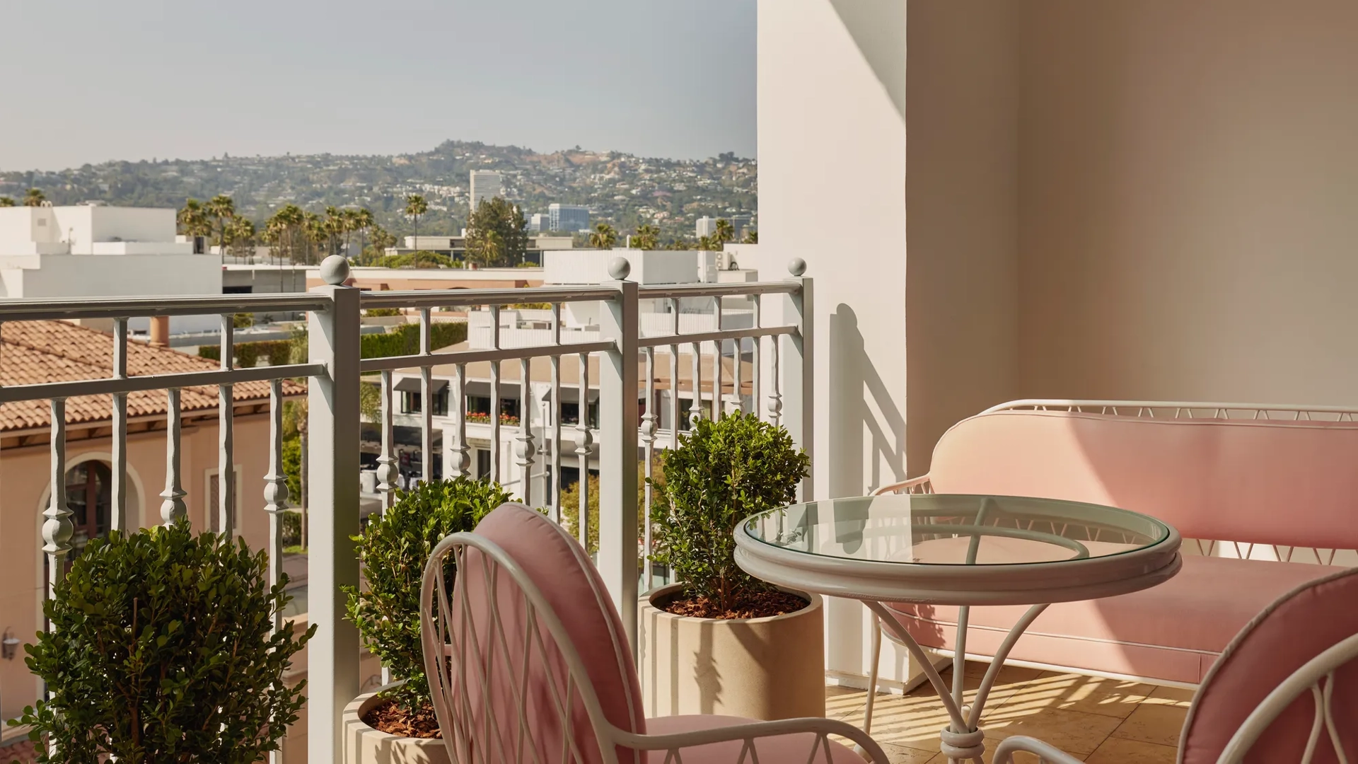 Balcony view with pink cushioned chairs and sofa, glass-topped table, and potted greenery, overlooking terracotta rooftops and distant hills under a bright sky.