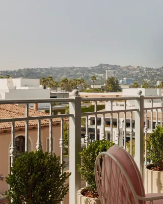Balcony view with pink cushioned chairs and sofa, glass-topped table, and potted greenery, overlooking terracotta rooftops and distant hills under a bright sky.