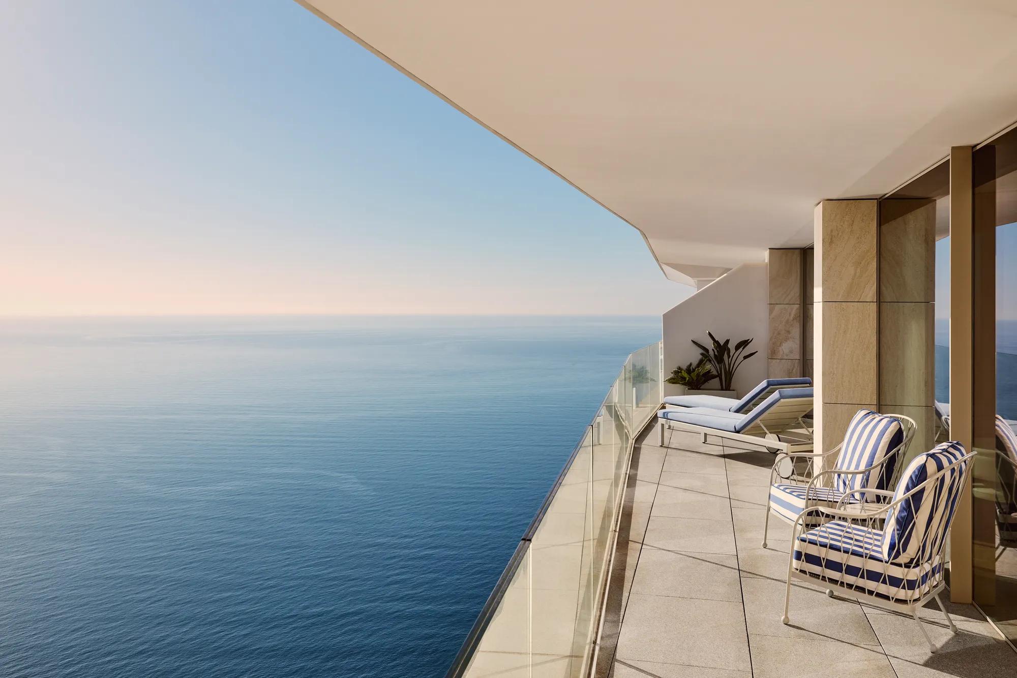 Spacious balcony with striped chairs, sun loungers, and glass railing, offering panoramic sea views beneath a clear sky.