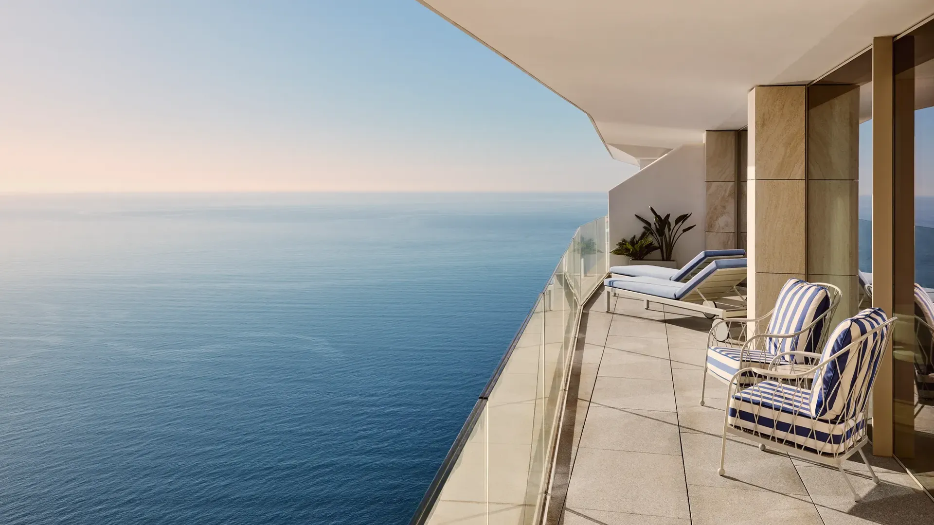 Spacious balcony with striped chairs, sun loungers, and glass railing, offering panoramic sea views beneath a clear sky.