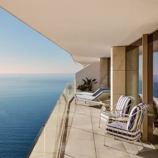 Balcon spacieux avec fauteuils rayés, transats et garde-corps en verre, offrant une vue panoramique sur la mer sous un ciel dégagé.