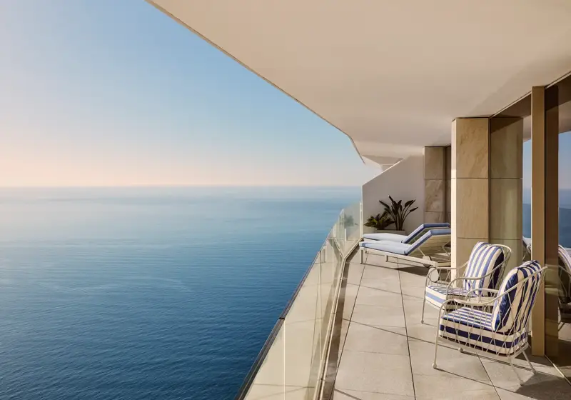 Spacious balcony with striped chairs, sun loungers, and glass railing, offering panoramic sea views beneath a clear sky.