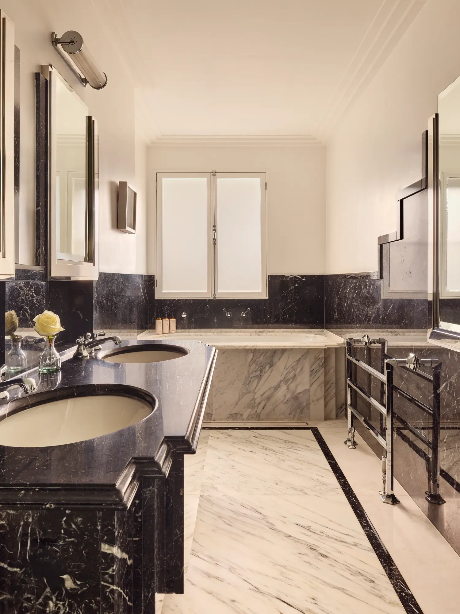 Luxurious marble bathroom featuring dual sinks with dark stone countertops, a built-in bathtub with light marble surround, and chrome fixtures under soft natural light from a frosted window.