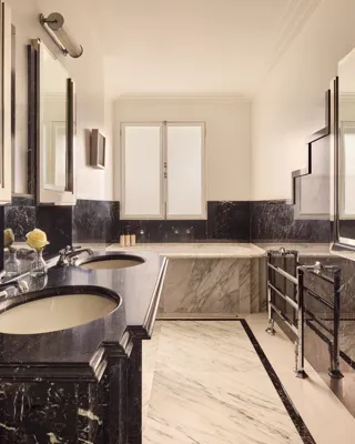Luxurious marble bathroom featuring dual sinks with dark stone countertops, a built-in bathtub with light marble surround, and chrome fixtures under soft natural light from a frosted window.