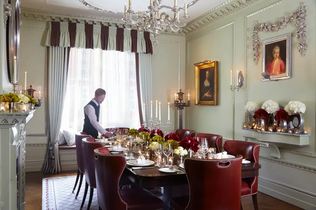 A butler sets a candlelit dining table with red leather chairs, floral centrepieces and ornate décor in an elegant private room.