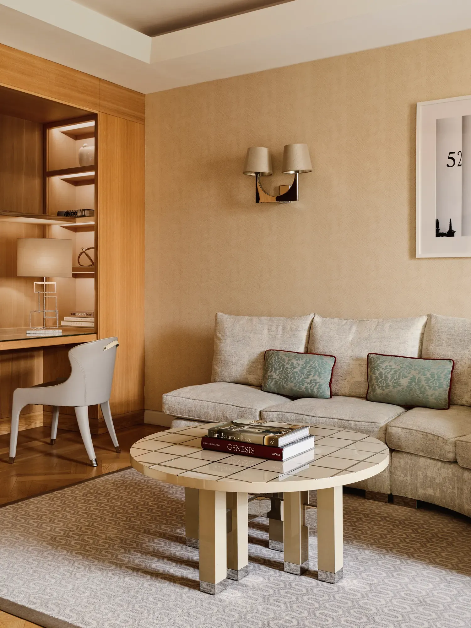 Bright suite corner with a cream sectional sofa, tiled coffee table, built-in wooden desk with chair and shelving, and wall sconces in a warm-toned interior.