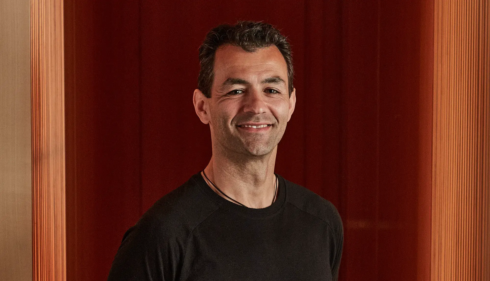 Smiling man in black T-shirt standing against rich red panelled wall.