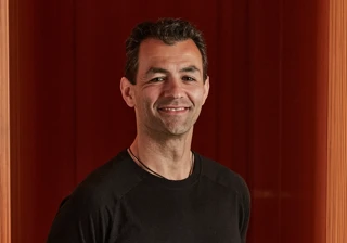 Smiling man in black T-shirt standing against rich red panelled wall.