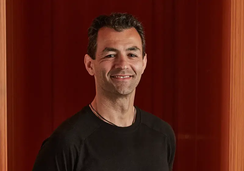 Smiling man in black T-shirt standing against rich red panelled wall.