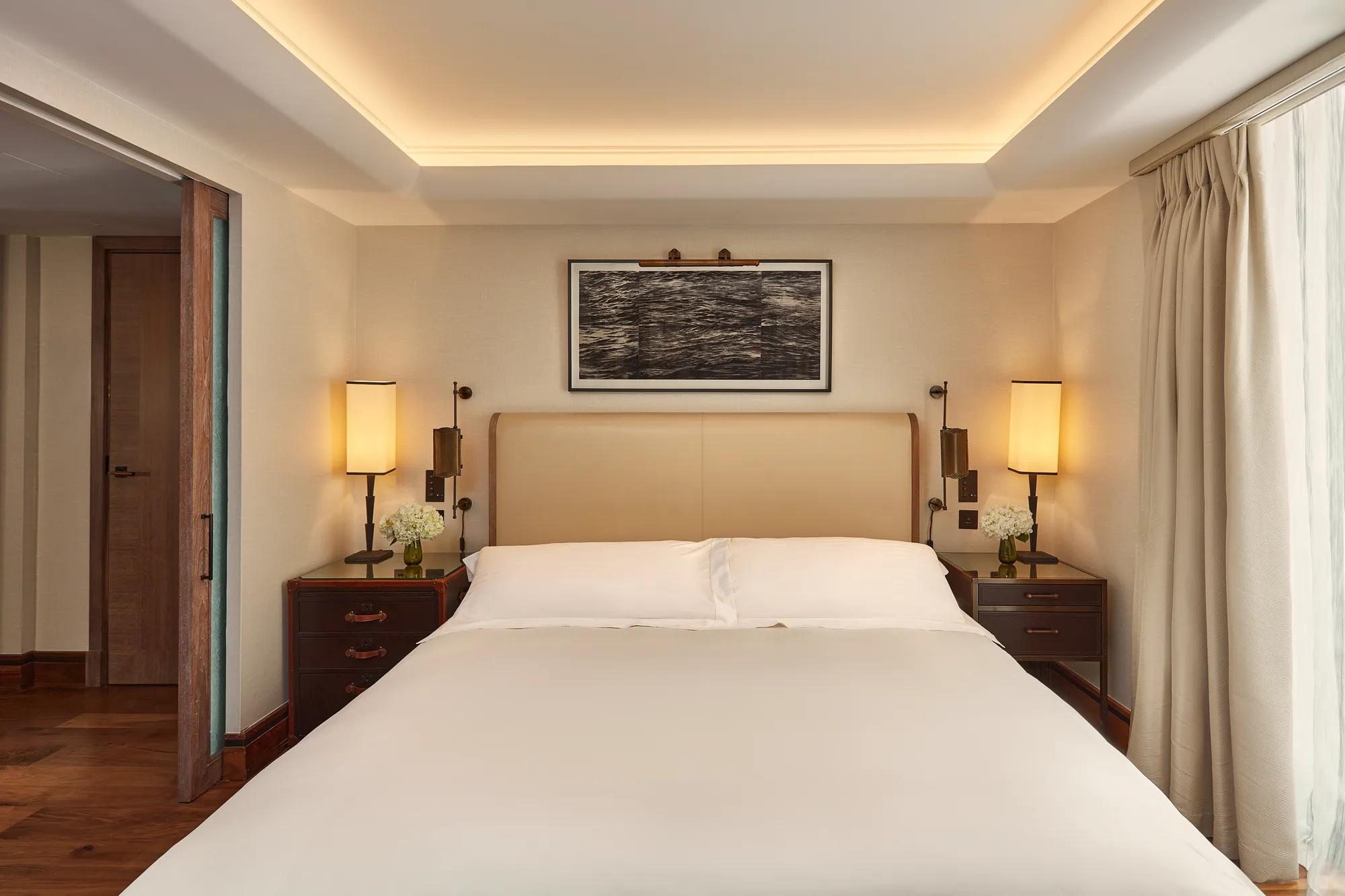 Calm bedroom with a cream headboard, bedside lamps, wooden nightstands with flowers, and light curtains framing a window.