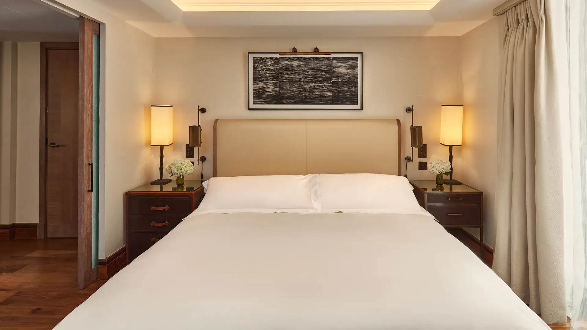 Calm bedroom with a cream headboard, bedside lamps, wooden nightstands with flowers, and light curtains framing a window.