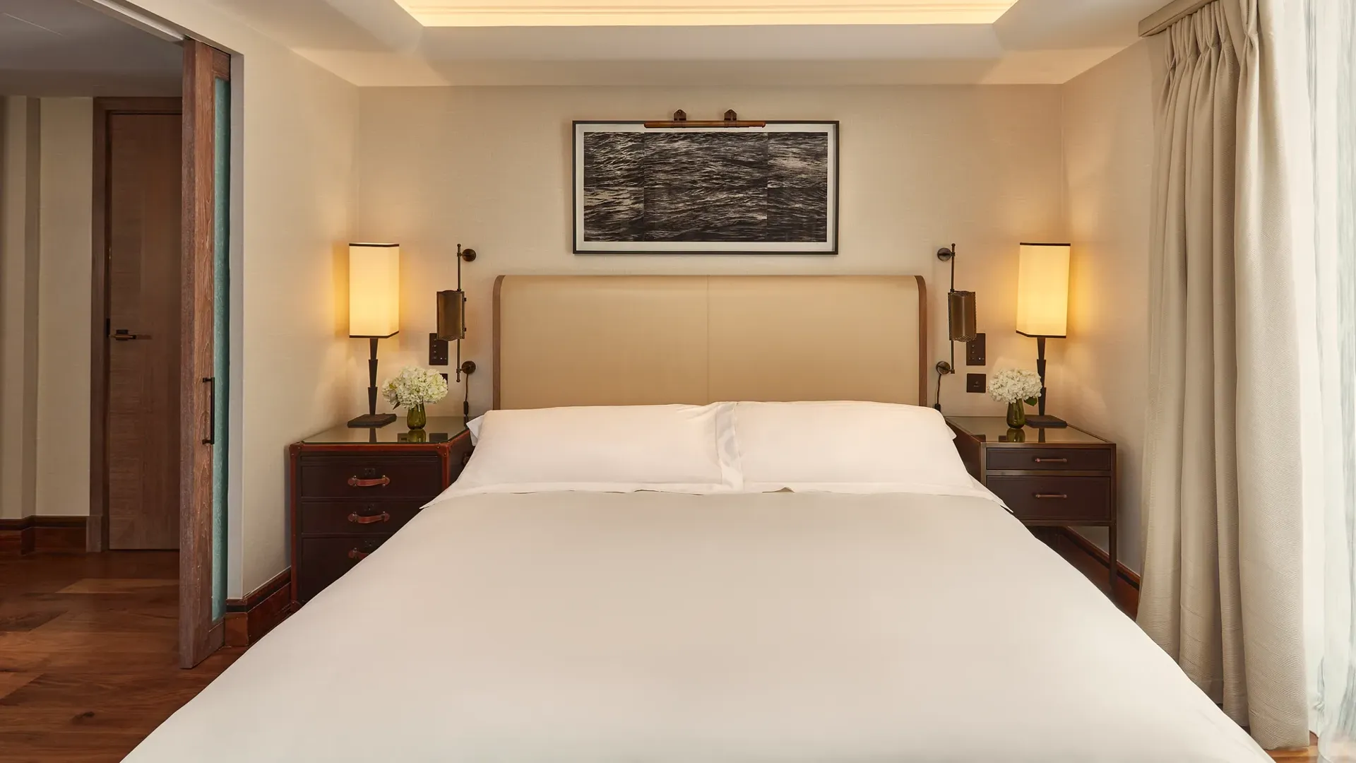 Calm bedroom with a cream headboard, bedside lamps, wooden nightstands with flowers, and light curtains framing a window.