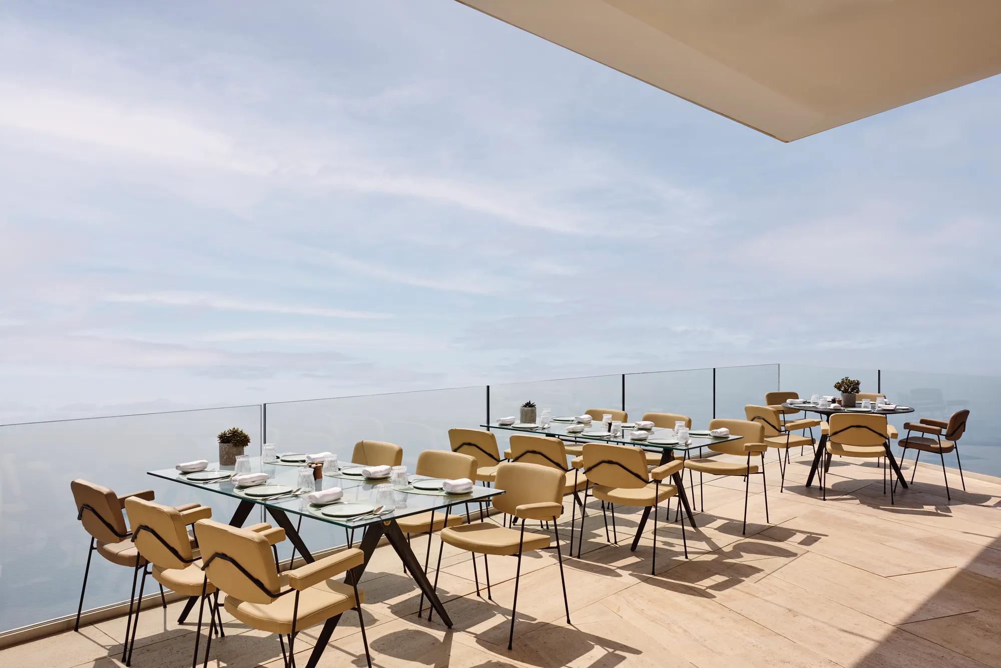Espace de restauration en terrasse avec des tables en verre modernes et des chaises beiges, offrant une vue dégagée sur la mer et le ciel.