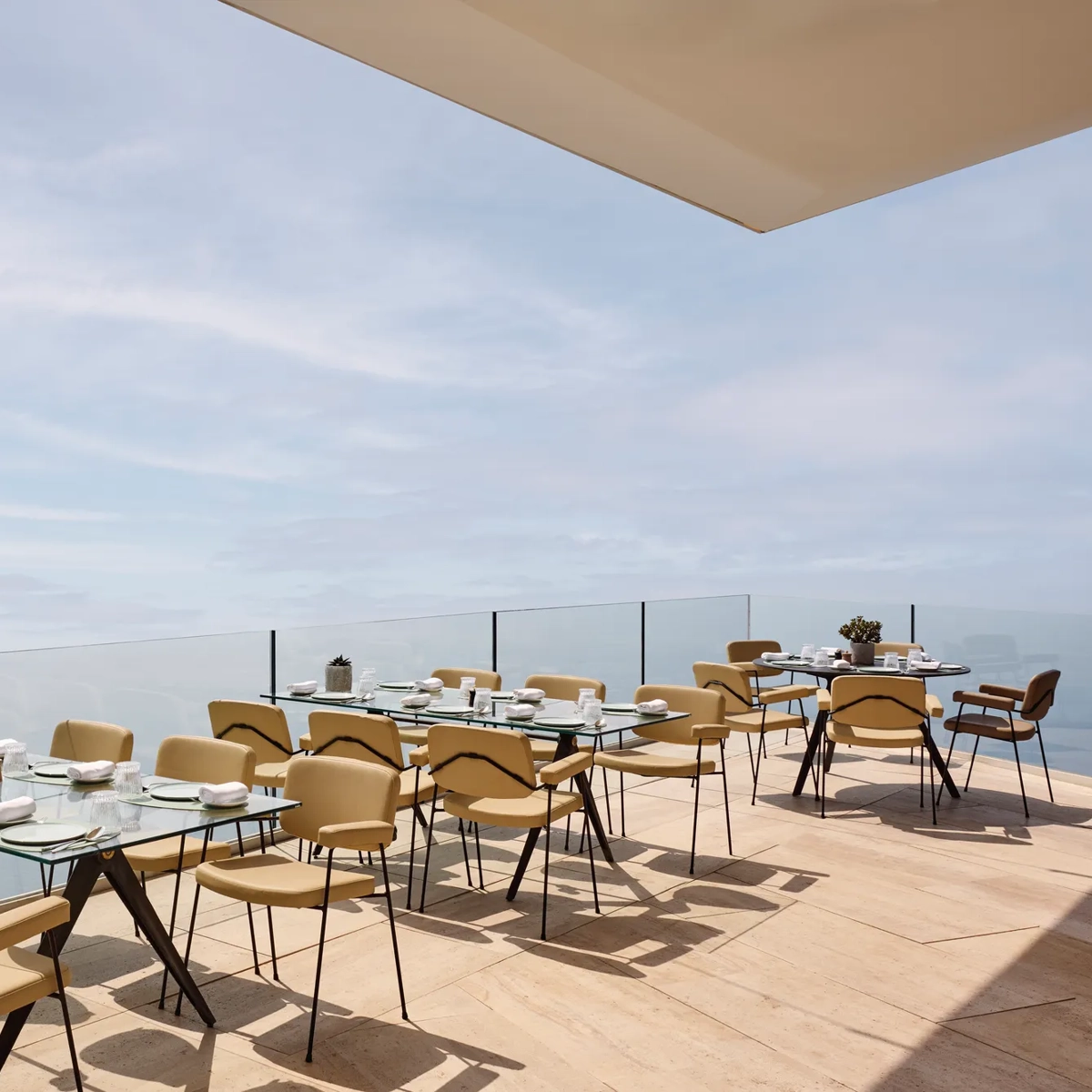 Espace de restauration en terrasse avec des tables en verre modernes et des chaises beiges, offrant une vue dégagée sur la mer et le ciel.