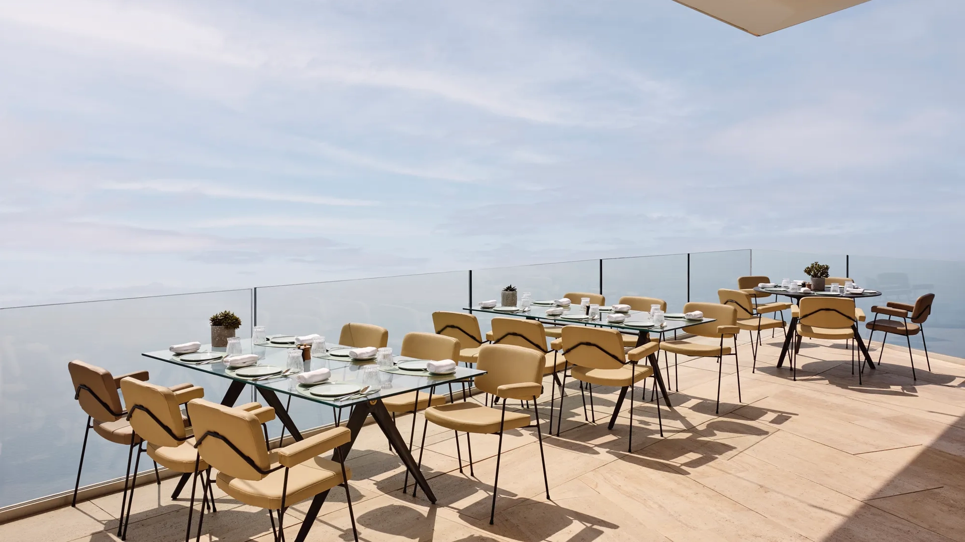 Espace de restauration en terrasse avec des tables en verre modernes et des chaises beiges, offrant une vue dégagée sur la mer et le ciel.