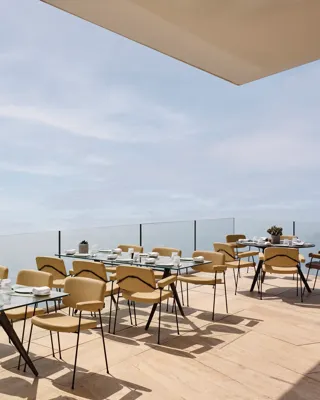 Terrace dining area with modern glass tables and beige chairs, set against a backdrop of the open sea and sky.