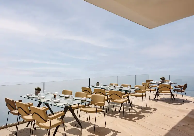 Espace de restauration en terrasse avec des tables en verre modernes et des chaises beiges, offrant une vue dégagée sur la mer et le ciel.