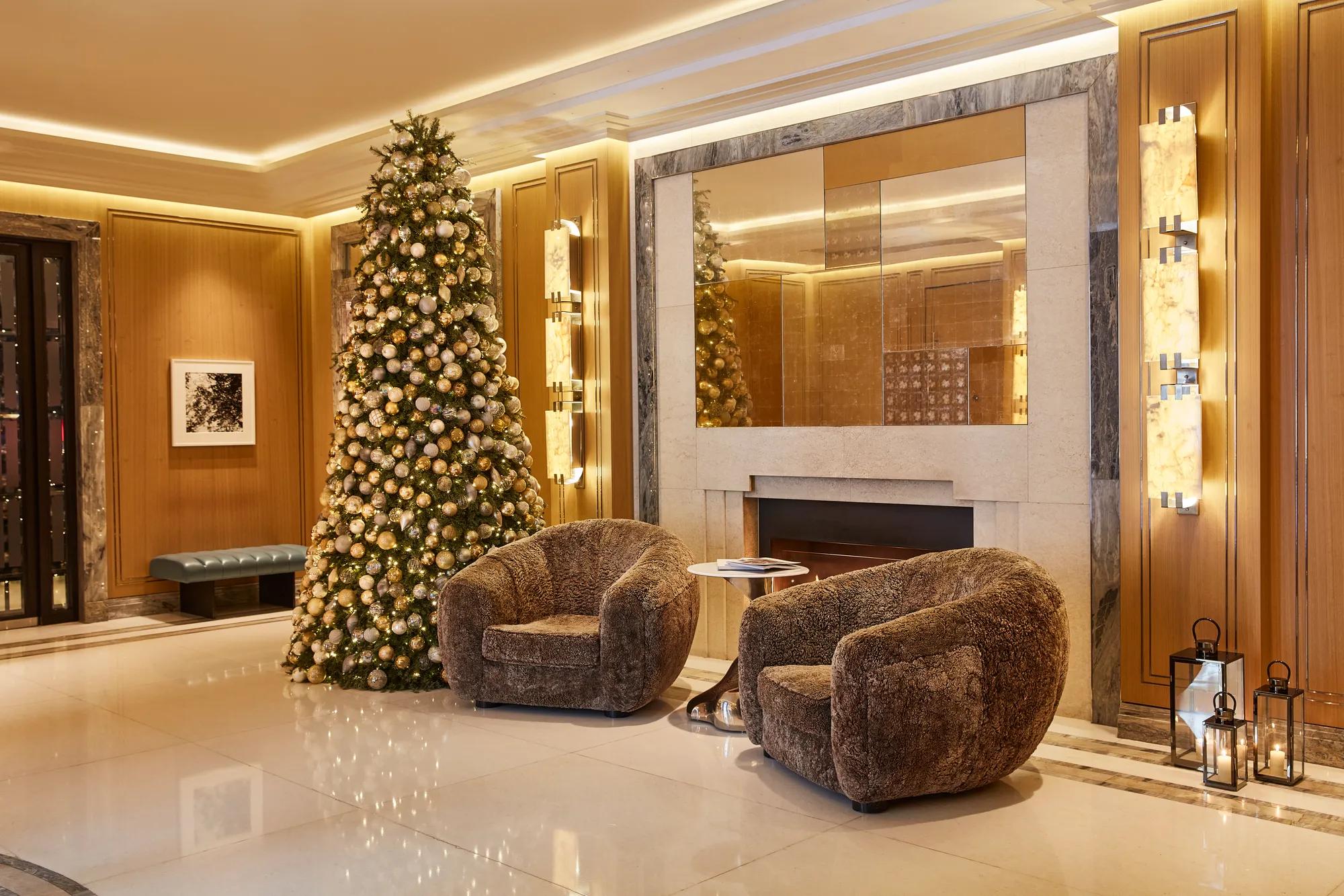 Elegant hotel lobby decorated for the festive season, featuring a tall Christmas tree adorned with gold and silver baubles beside two plush armchairs, set against a marble fireplace and warm wood-panelled walls illuminated by soft ambient lighting.