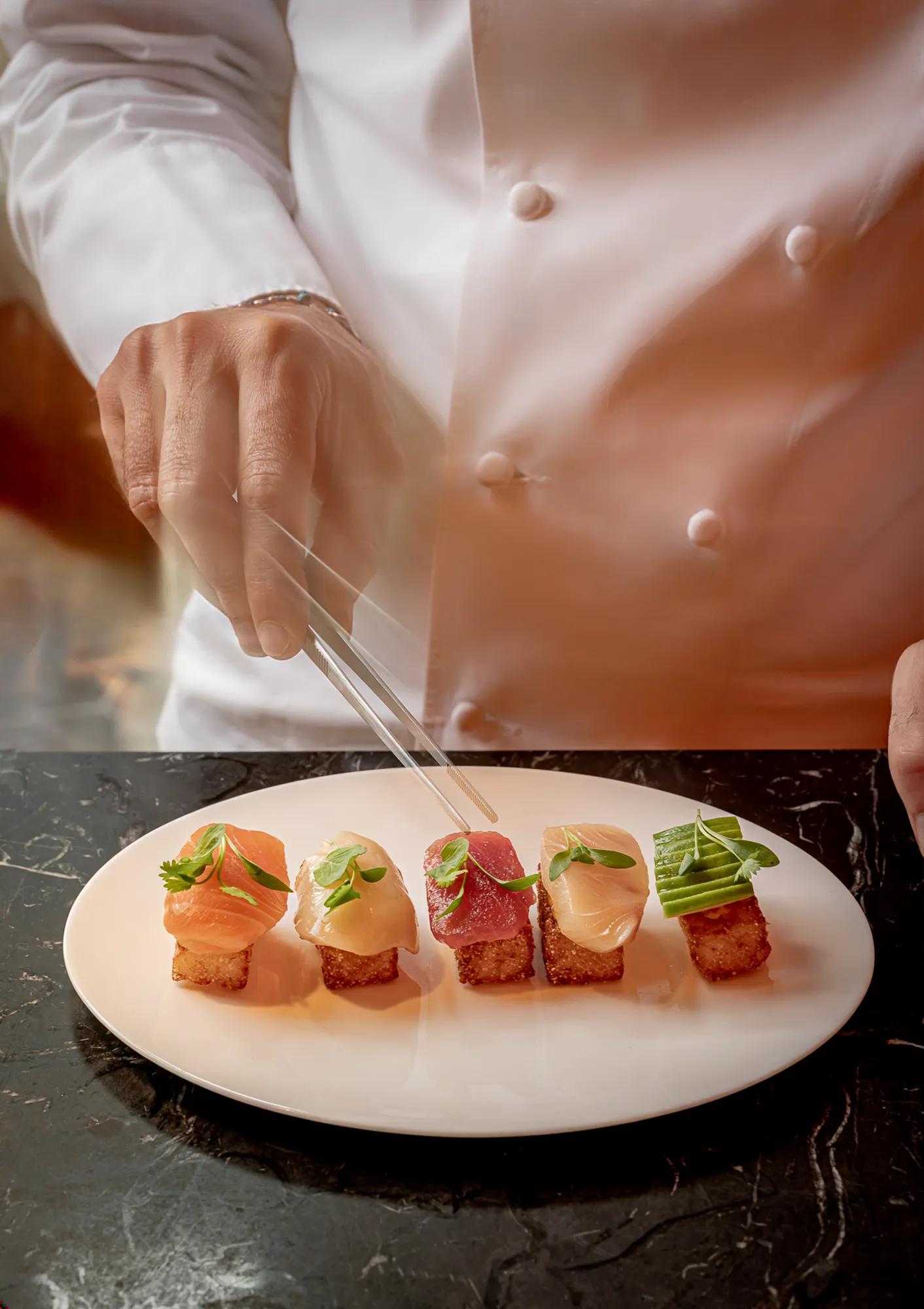 Chef arranging sushi canapés on a white plate using tweezers at a marble counter.
