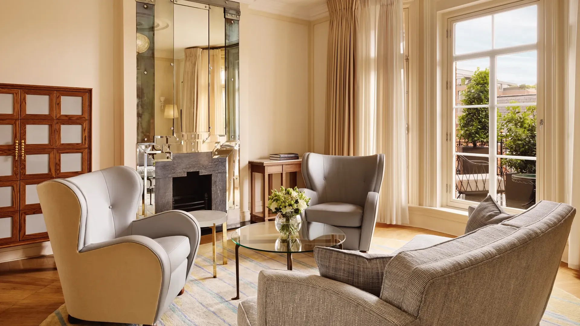 Elegant sitting area with grey armchairs and a matching sofa around a glass coffee table topped with flowers, set beside large windows and a mirrored fireplace reflecting warm natural light.