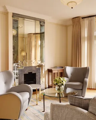 Elegant sitting area with grey armchairs and a matching sofa around a glass coffee table topped with flowers, set beside large windows and a mirrored fireplace reflecting warm natural light.