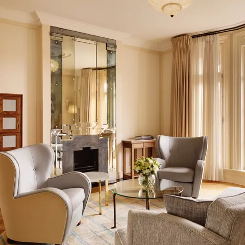 Elegant sitting area with grey armchairs and a matching sofa around a glass coffee table topped with flowers, set beside large windows and a mirrored fireplace reflecting warm natural light.