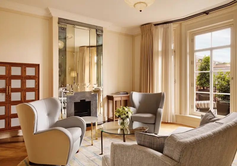 Elegant sitting area with grey armchairs and a matching sofa around a glass coffee table topped with flowers, set beside large windows and a mirrored fireplace reflecting warm natural light.