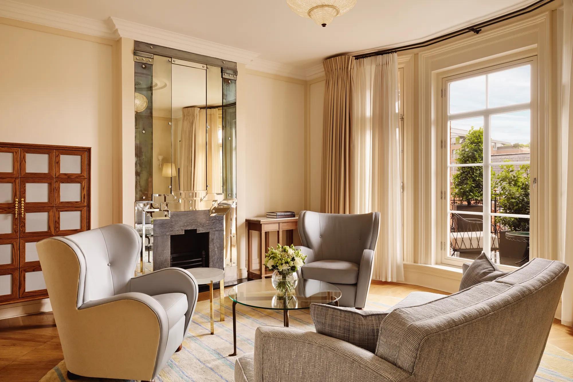 Elegant sitting area with grey armchairs and a matching sofa around a glass coffee table topped with flowers, set beside large windows and a mirrored fireplace reflecting warm natural light.