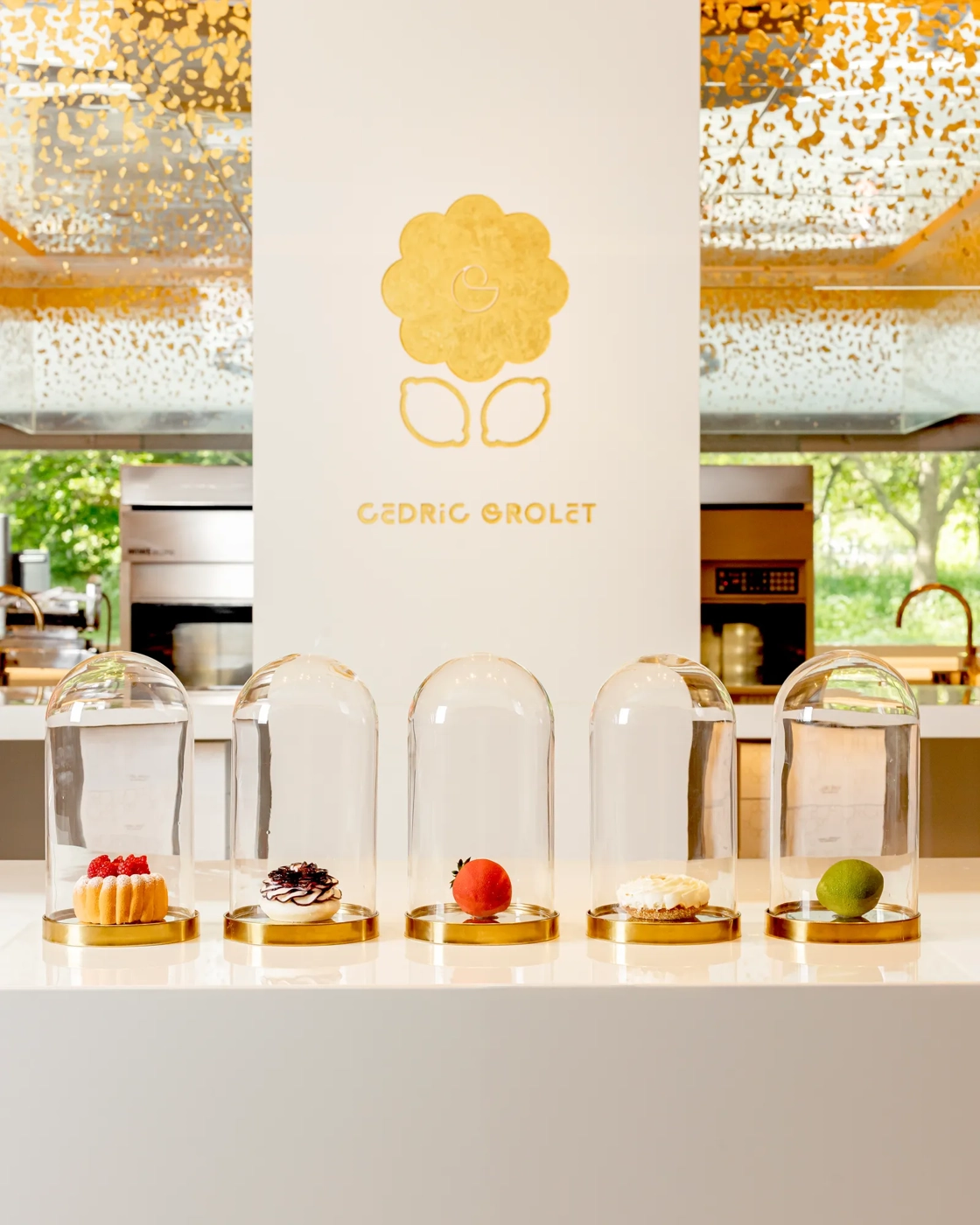 Five intricate pastries displayed under glass domes at Cédric Grolet patisserie, with gold detailing and modern backdrop.