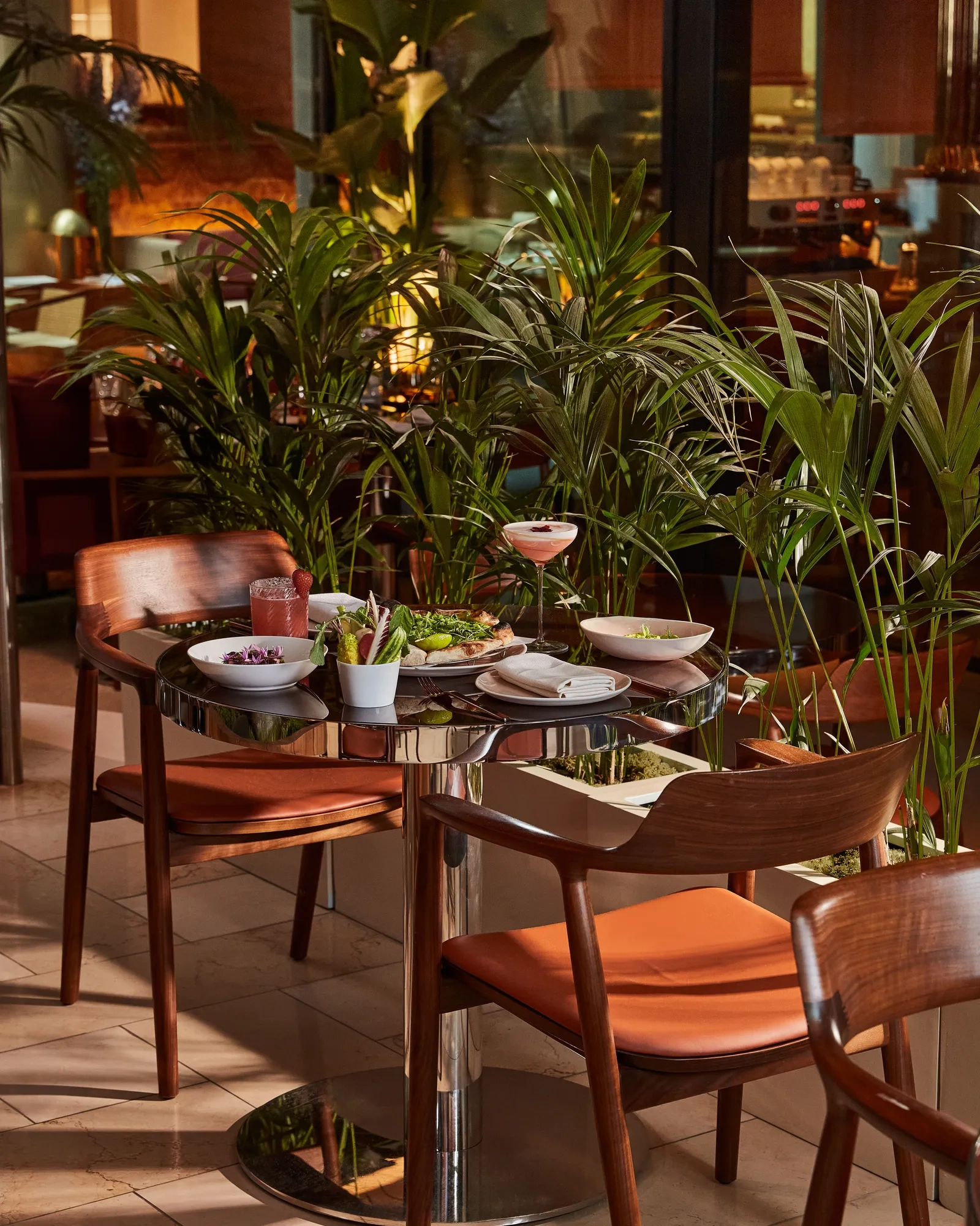 Dining table for two with cocktails and sharing plates, surrounded by potted palms and warm wood furnishings.