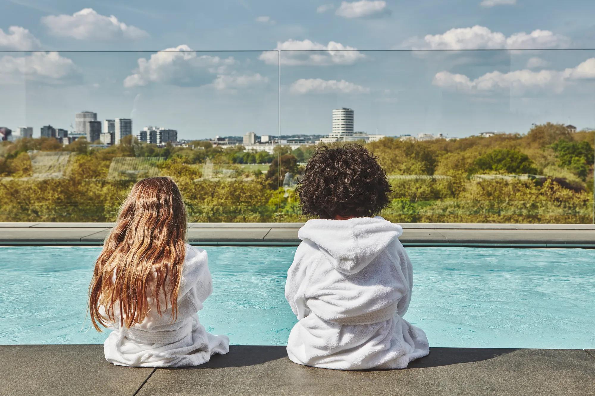 Two children in white robes sit by a rooftop pool, facing a leafy city skyline under a blue, cloud-dotted sky.