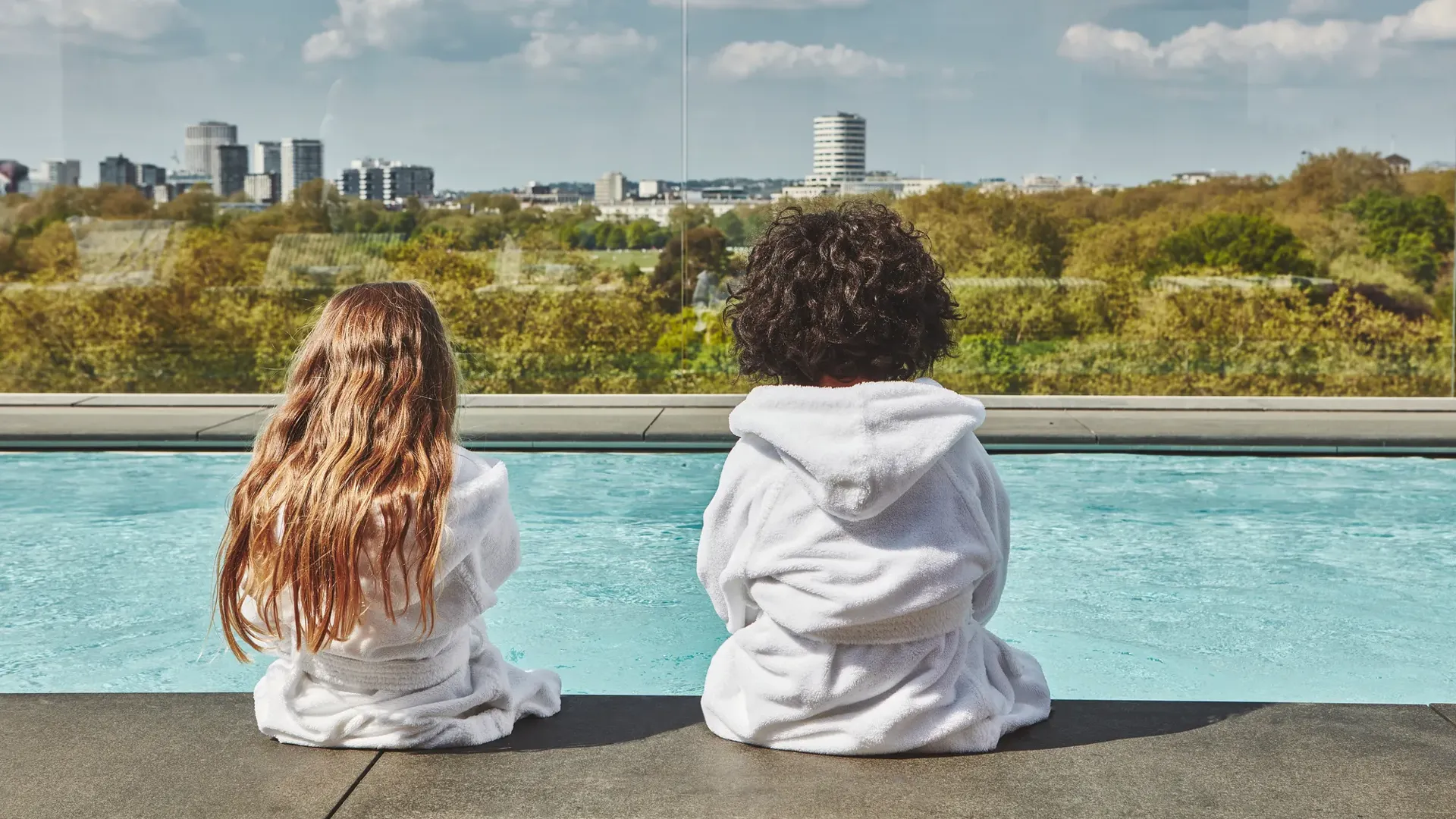 Two children in white robes sit by a rooftop pool, facing a leafy city skyline under a blue, cloud-dotted sky.