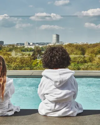 Two children in white robes sit by a rooftop pool, facing a leafy city skyline under a blue, cloud-dotted sky.