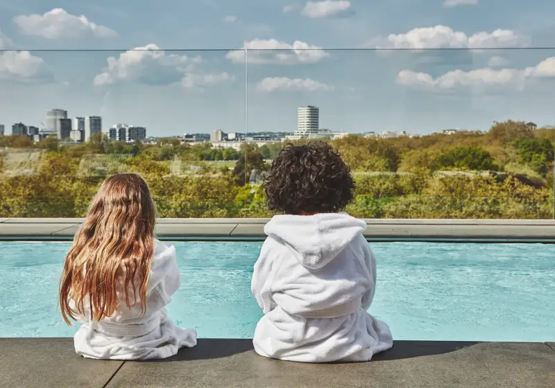 Two children in white robes sit by a rooftop pool, facing a leafy city skyline under a blue, cloud-dotted sky.