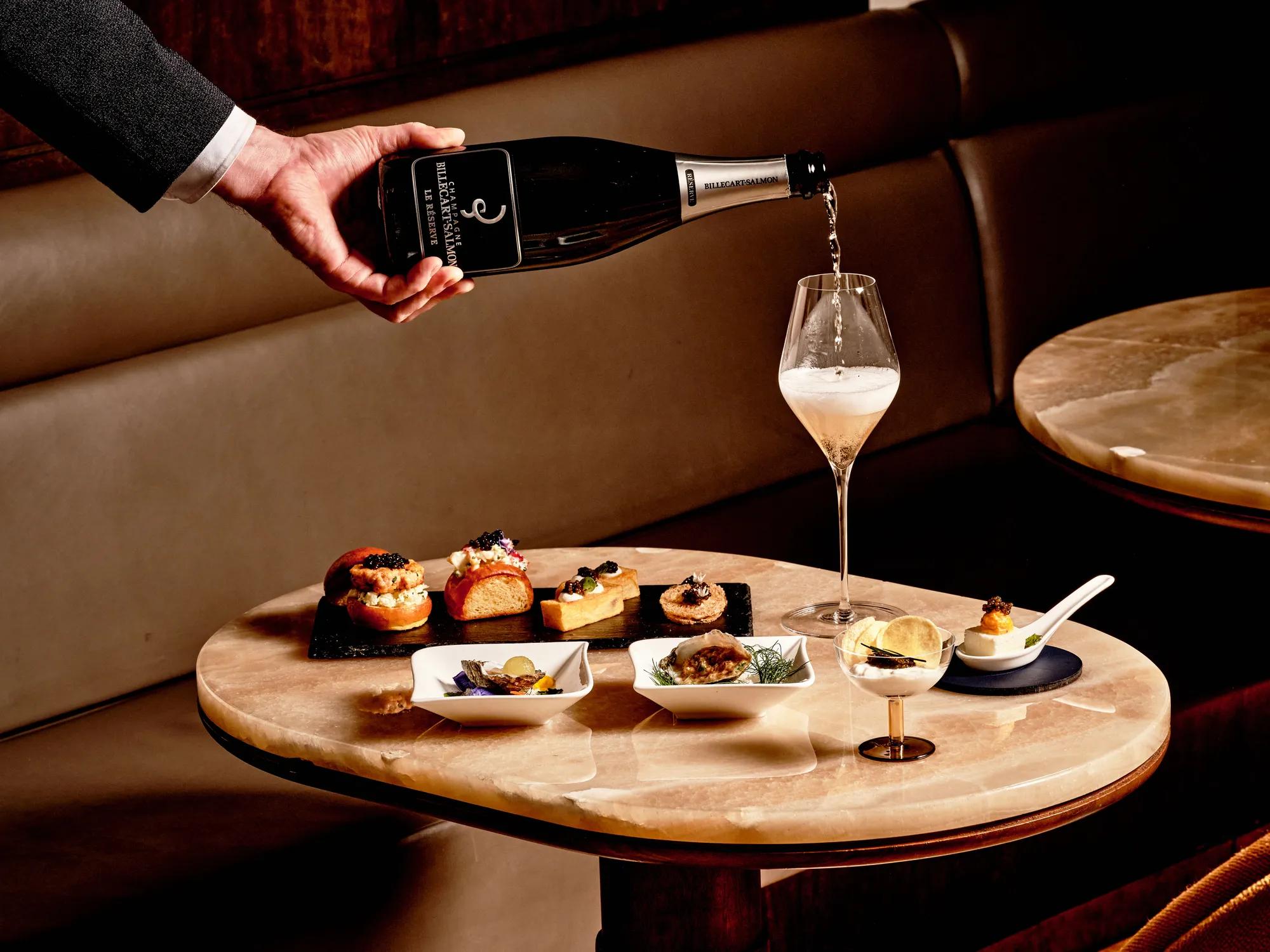 Hand pouring champagne into flute beside gourmet canapés and caviar at Maybourne Beverly Hills