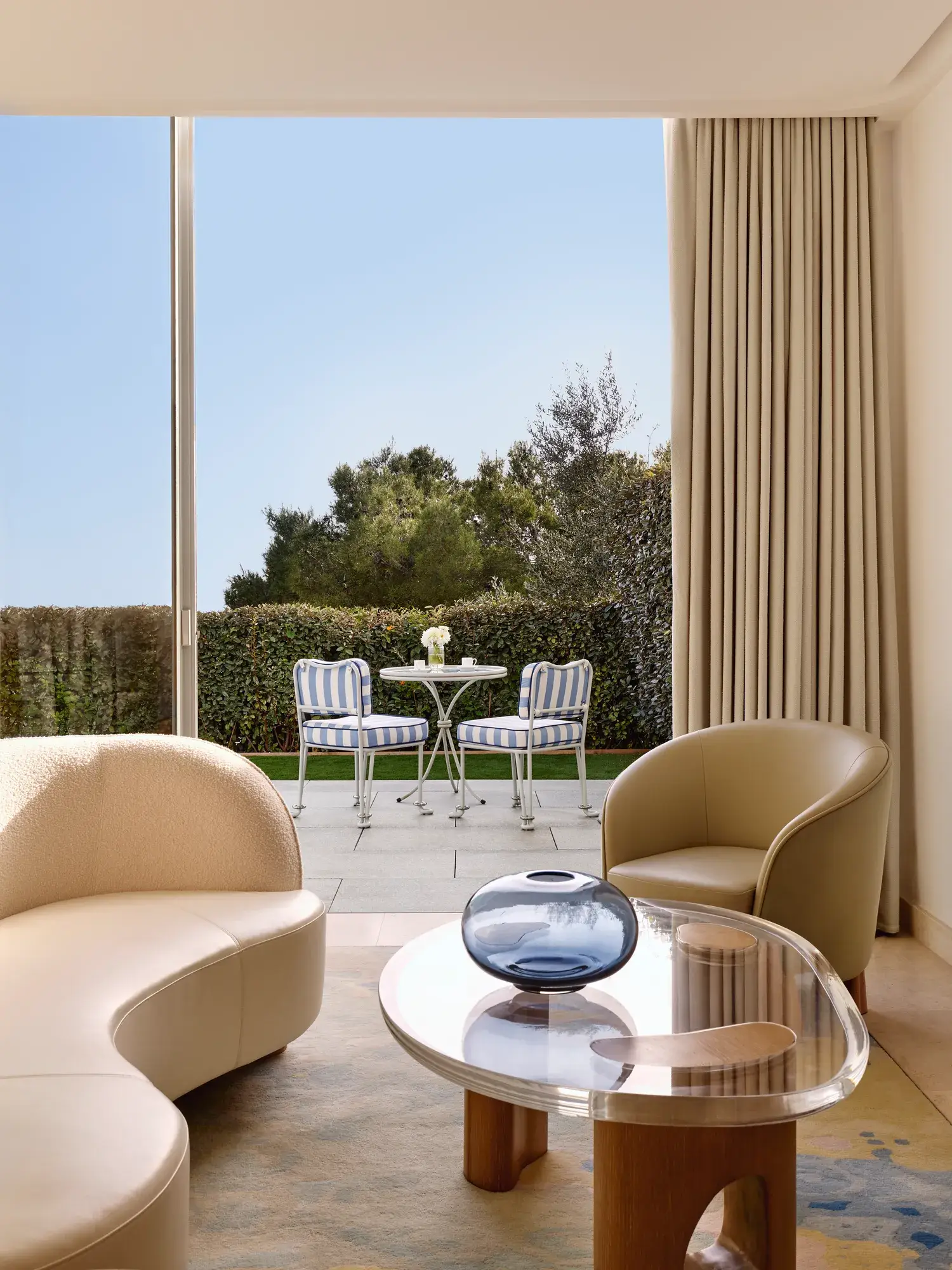 Coin salon élégant avec un canapé incurvé, un fauteuil moderne et une table basse en verre donnant sur une terrasse privée avec des chaises rayées et une table, le tout entouré de verdure.